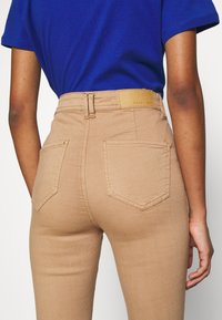 Beige högmidjade jeans med en slät yta, som har två bakfickor och en brun etikett vid midjan. Kontrastsydd synlig.