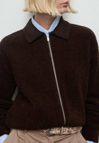 Bruine fluweelachtige zip-up jas met een donkere kraag, voorzien van een zilveren rits en een lichtblauwe shirtkraag eronder, gecombineerd met een gemusterde riem.