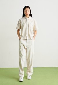 Two-piece outfit in light beige with vertical stripes, featuring a short-sleeve button-up shirt and relaxed-fit pants. White sneakers worn.