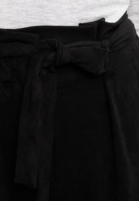 Černý textilní pásek s mašlí, texturovaný povrch a hladký finiš. Uvázaný kolem oblečení s šedým topem.