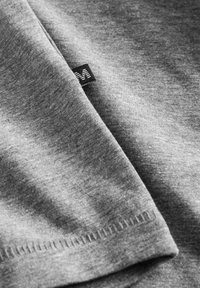 Tejido gris con una textura suave, que presenta un borde doblado y una pequeña etiqueta negra que muestra un logotipo en blanco. Costuras bien hechas visibles.