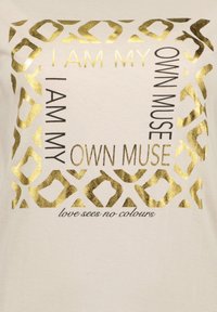 Vaaleanbeige t-paita, jossa on kultainen metallinen leopardiprinttiraita. Tekstissä lukee "Olen oma muusani" ja "rakkaus ei näe värejä" mustalla letteringillä.