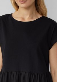 Frau in einem schwarzen Kurzarmkleid mit rundem Ausschnitt und geraffter Taille, bis zur Mitte der Taille zugeschnitten.