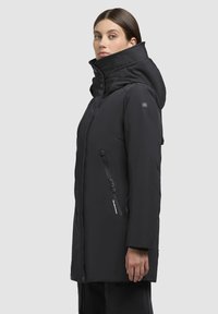 Frau in einem schwarzen Wintermantel mit Kapuze, hohem Kragen und mit Reißverschlüssen versehenen Taschen, steht seitlich vor einem schlichten weißen Hintergrund.