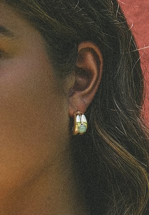 Goldener Creolen-Ohrring mit kleinem rundem grünem Stein, getragen am Ohr einer Person mit dunklem Haar vor rotem Hintergrund.