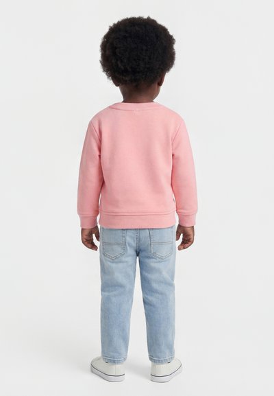 Enfant aux cheveux bouclés foncés portant un sweat-shirt rose, un jean bleu clair et des baskets blanches, de dos sur fond blanc.