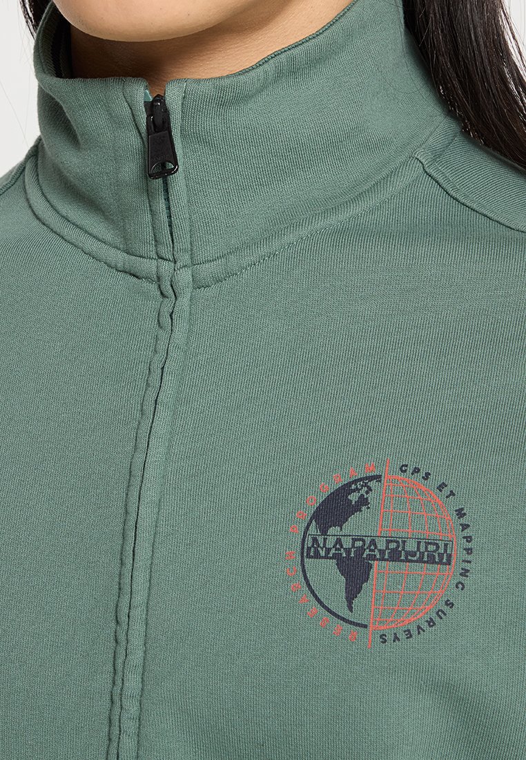Giacca in pile verde con colletto alto, zip nera e logo circolare in rosso e blu scuro con un design del globo. Testo attorno al logo.