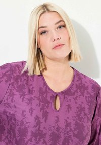 Lila Oberteil mit strukturiertem Muster, rundem Ausschnitt mit kleinem Schlüssellochdetail und kurzen Ärmeln. Weiches Material mit leichtem Glanz.