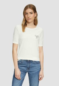 Witte t-shirt met korte mouwen van soepel materiaal, met een klein zwart strikdesign op de linkerkant van de borst. Spijkerbroek gedeeltelijk zichtbaar.