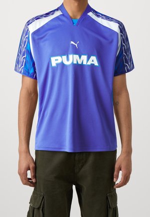 Mann trägt ein lila Puma-Sporttrikot mit weißen und blauen Akzenten und dunkelgrünen Cargohosen, steht vor einem schlichten Hintergrund.