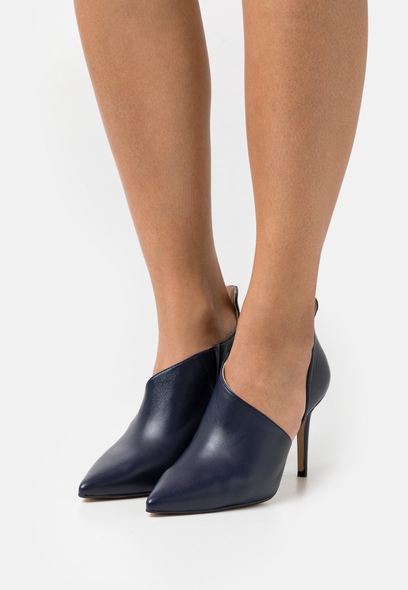 Escarpins en cuir bleu marine à bouts pointus portés pieds nus, avec un design élégant et minimaliste et des talons aiguilles.
