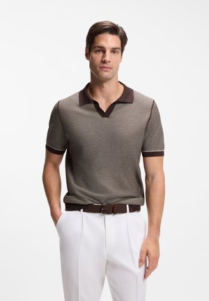 Uomo che indossa una polo marrone a maniche corte in maglia con colletto scuro e pantaloni bianchi plissettati, in piedi con le mani nelle tasche contro uno sfondo bianco.