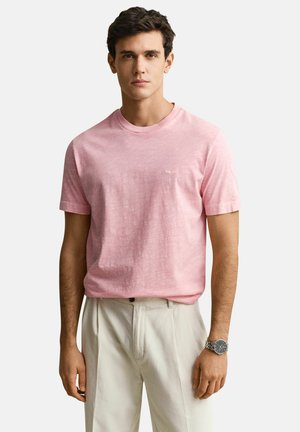 Jonge man die een lichtroze T-shirt met korte mouwen, beige broek en een zilveren polshorloge draagt, staand tegen een effen achtergrond.