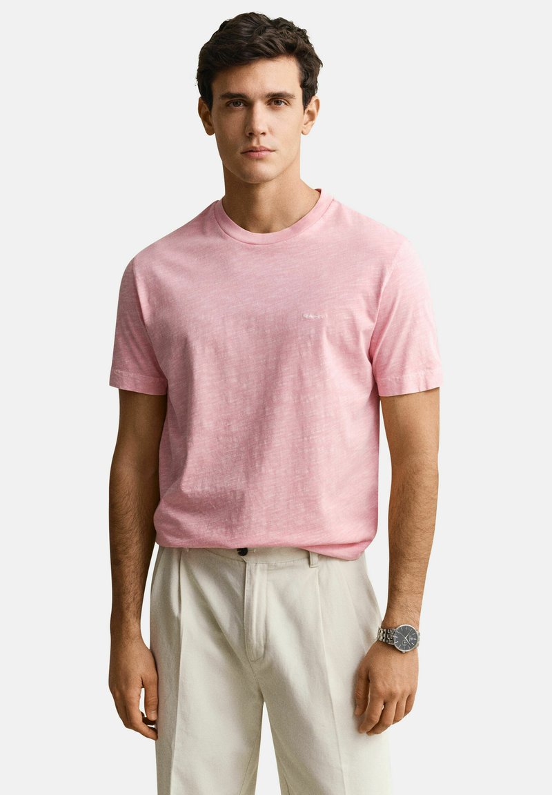 Jeune homme portant un t-shirt à manches courtes rose clair, un pantalon beige et une montre-bracelet en argent, debout devant un fond uni.