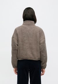 Bluza polarowa w stonowanym brązie, z wysokim kołnierzem, opuszczonymi ramionami i prążkowaniem. Miękka, teksturowana tkanina o luźnym kroju.