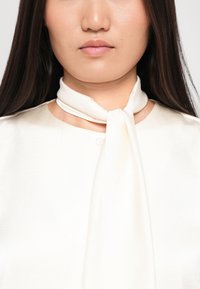Bluză albă cu guler tip fundă, confecționată dintr-un material fin, cu un detaliu cu buton la gât și umeri fără cusături.