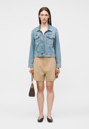 Γυναίκα που φοράει τζιν μπουφάν γαλάζιου χρώματος, μπεζ σορτς, μαύρα παπούτσια, κρατώντας μια μικρή καφέ τσάντα πάνω σε απλό φωτεινό φόντο.