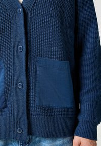 Granatowy sweter zapinany na guziki z włóczki, o teksturowanym wzorze, z dwiema gładkimi kieszeniami, okrągłym dekoltem i sześcioma guzikami z przodu.
