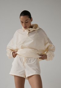 Lekki beżowy sweter z wysokim kołnierzem i elastycznymi mankietami, w zestawie z pasującymi szortami z elastycznym pasem. Gładkie, matowe wykończenie.