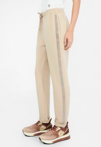 Pantalons de jogging beiges avec une taille à cordon, dotés d'une bande argentée brillante sur le côté. Associés à des chaussures de sport marron.