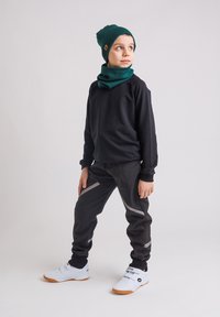 Chłopiec w ciemnym stroju sportowym, zielonej czapce i szaliku, stojący z jedną nogą do przodu, zwrócony w prawo na tle jednolitego koloru.