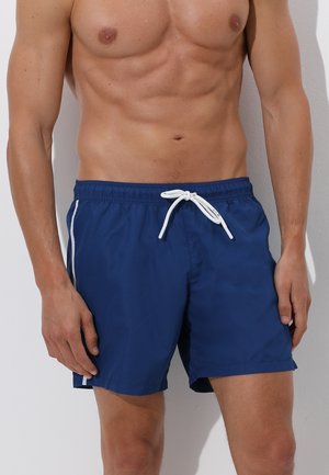 Torso maschile che indossa pantaloncini da bagno blu con coulisse bianca e strisce laterali, in piedi contro uno sfondo chiaro.