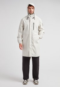 Impermeabile, giacca leggera grigia con cappuccio, polsini elasticizzati, tasca frontale con zip e cordino regolabile al colletto. Pantaloni e scarpe neri.
