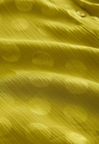 Gelbe Bluse aus einem strukturierten Stoff mit großen, erhobenen Punkten. Sichtbares Knopfdetail und eine glatte, leicht glänzende Oberfläche.