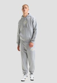 Set di felpa grigia con cappuccio e pantaloni della tuta in pile; presenta una tasca frontale, cappuccio con cordino e dettagli del logo in crema. Abbinato a sneakers.