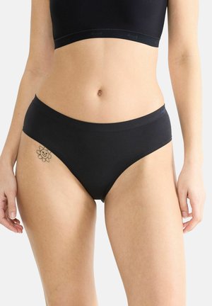 Eine Frau trägt nahtlose schwarze Unterwäsche und einen passenden schwarzen Bralette, auf der linken Hüfte ist ein kleines lächelndes Blümchen-Tattoo zu sehen.