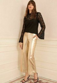 Top lungo in pizzo nero con maniche a campana, abbinato a pantaloni larghi dorati lucidi e tacchi a strappo neri.