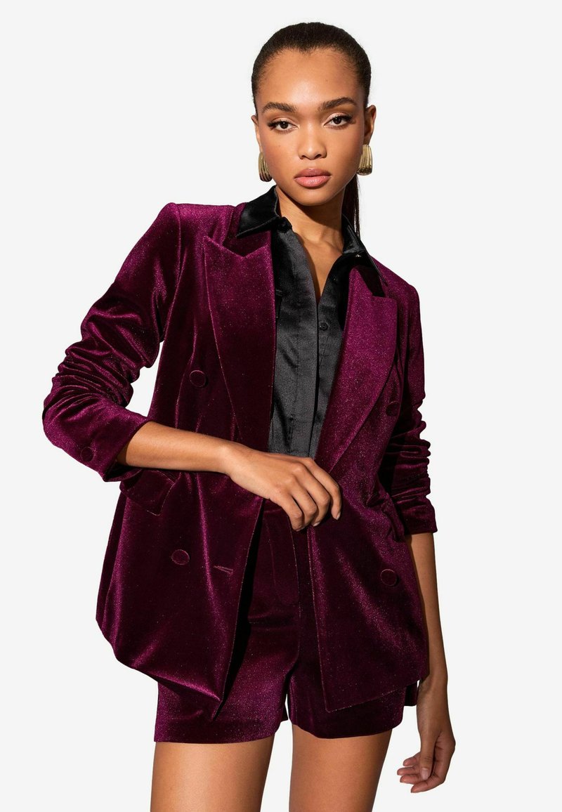 Bordó bársonyből készült blézer dupla gombolással, fekete inggel és rövidnadrággal párosítva. Simább textúra diszkrét fényeléssel és strukturált sziluettel.