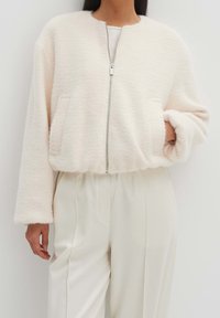 Chaqueta recortada de color crema y textura suave, con cierre de cremallera, dos bolsillos frontales y mangas de corte holgado.