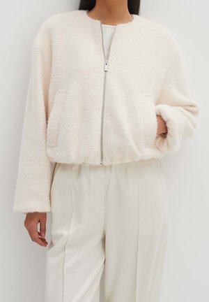 Veste courte floue de couleur crème avec fermeture éclair, deux poches avant et manches amples, fabriquée à partir d'un tissu doux et texturé.