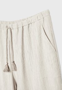 Pantalon en tissu beige texturé avec taille élastique, cordon ajustable avec pompons et poche latérale droite.