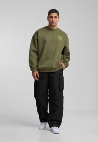 Olijfgroen sweatshirt met ronde hals en een groen grafisch logo, gecombineerd met zwarte cargobroek met meerdere zakken en witte sneakers.
