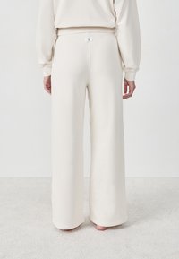Person, die weite, cremefarbene Sweatpants und ein passendes Oberteil trägt, barfuß auf einem hellen Teppich steht und mit dem Rücken zur Kamera blickt.