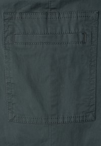 Poche arrière en tissu gris foncé avec un détail de poche passepoilée horizontale et des coutures visibles sur le vêtement.