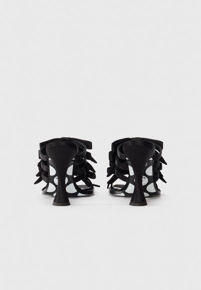 Une paire de sandales à talons hauts noirs avec des lanières, des talons incurvés et des pois blancs sur les semelles, vues de derrière.