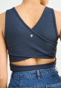 Navy blue cropped tank top with a textured fabric, wrap design, and a silver button closure at the back. Features a v-shaped back.