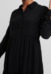 Robe longue noire à manches longues avec un col chemise, une patte de boutonnage et un design à volants ; fabriquée en tissu léger avec une texture lisse.