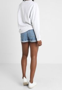 Osoba oblečená v svetle šedém mikině, zkrácených džínových šortkách a bílých teniskách, stojící s otočeným zády proti jednobarevnému bílému pozadí.