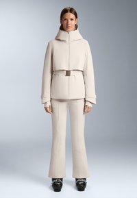 Beżowa krótka kurtka z wysokim kołnierzem i paskiem, w połączeniu z dopasowanymi spodniami w stylu dzwonów. Gładka, elegancka tekstura; czarne buty narciarskie z metalicznymi akcentami.