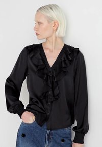 Blusa nera in satin con scollatura a V, davanti arricciato e maniche lunghe. Presenta chiusura a bottoni e polsini arricciati. Indossata con una gonna di jeans.