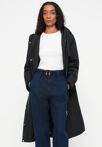 Zwarte lange parka met capuchon, gecombineerd met een wit T-shirt en hooggetailleerde blauwe broek met een riem en details in de zijnaad.