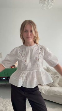 Jeune femme portant un chemisier blanc brodé et un pantalon noir, debout dans un salon avec un canapé et une armoire verte en arrière-plan.