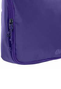 Bolsa de tela morada con superficie texturizada, cremallera negra y detalle de logotipo en relieve. Bordes lisos y fondo redondeado.