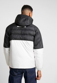 Giacca a vento con design a blocchi di colore bianco e nero, texture trapuntata e cappuccio. Include etichetta del marchio sul retro.