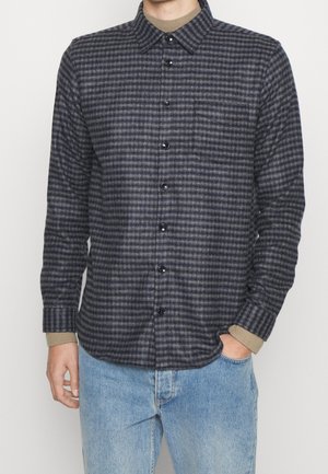 Chemise à carreaux bleu marine et gris en tissu doux, avec un devant boutonné, un col, une poche sur la poitrine et des accents de manches retroussées.