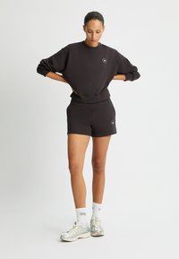 Maglione sportivo marrone scuro con scollo rotondo e polsini elasticizzati, abbinato a pantaloni corti a vita alta dello stesso colore, indossato con sneakers chiare.